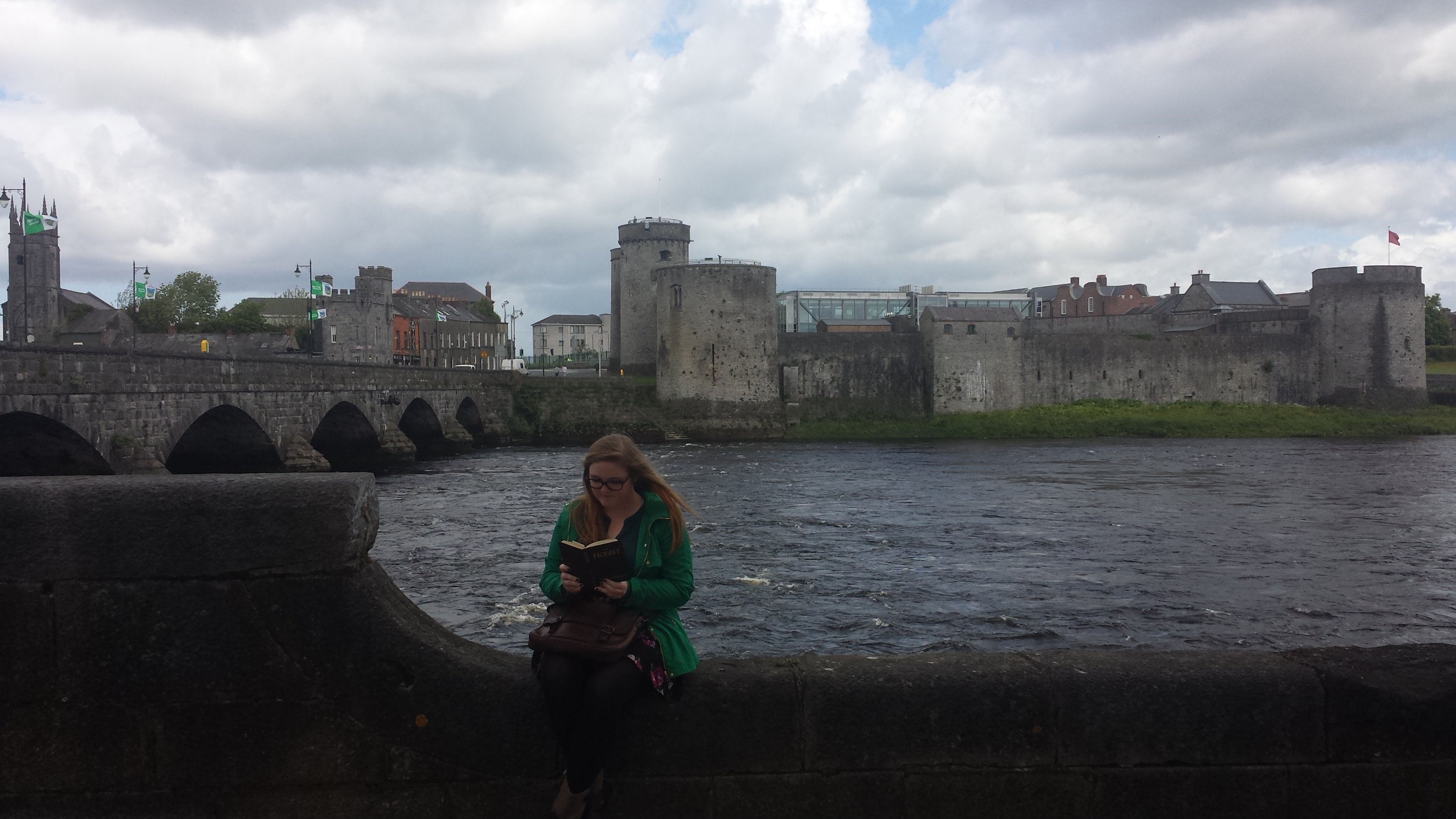 picture of King John's CAstle from the opposite side of the Shannon River. Bri sits on the ledge looking down at a book with the castle in the distance. The castle is more like stone walls with various towers.